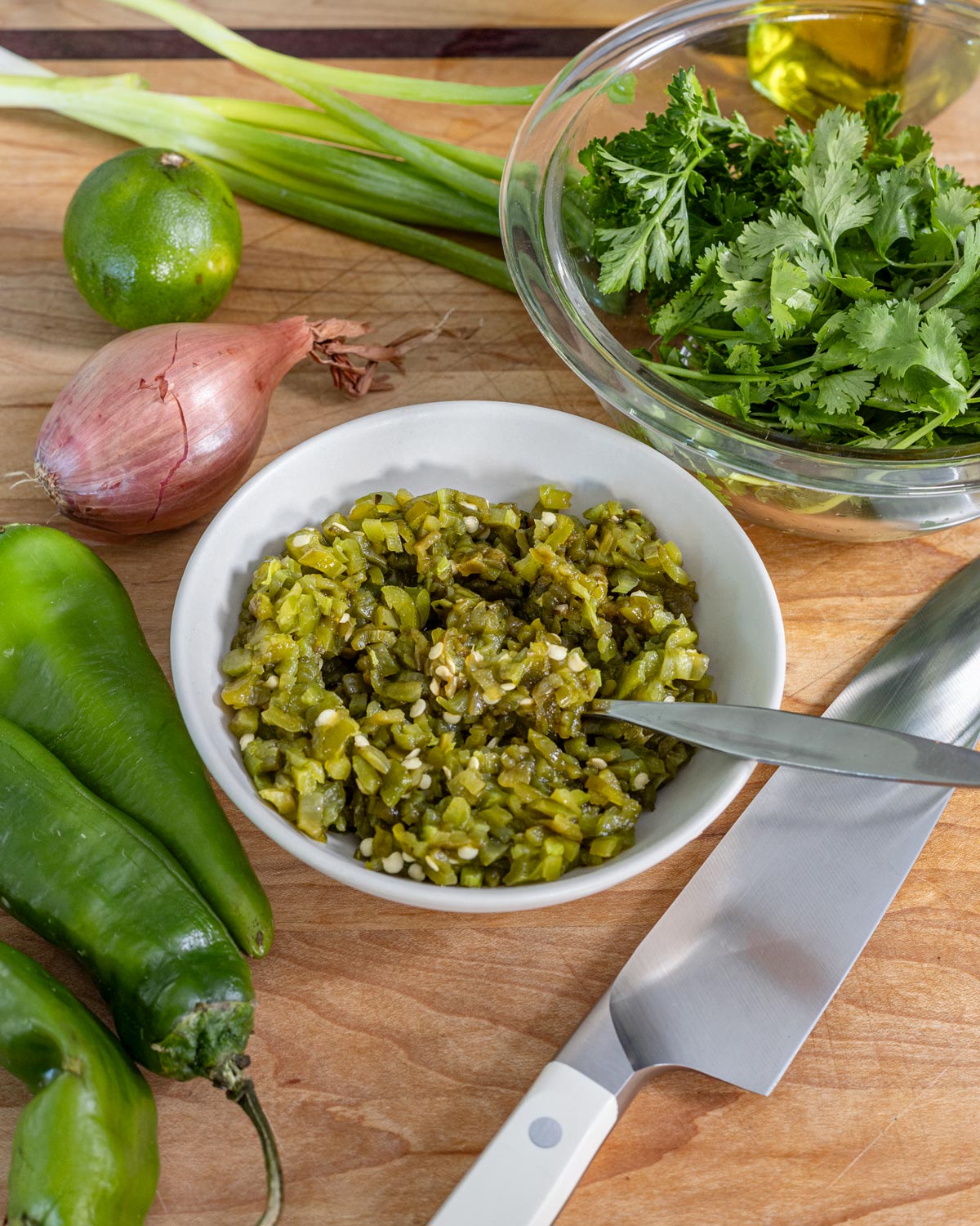 Chopped roasted green chiles in a dish with cilantro, parsley, scallions, lime, and a shallot on a cutting board