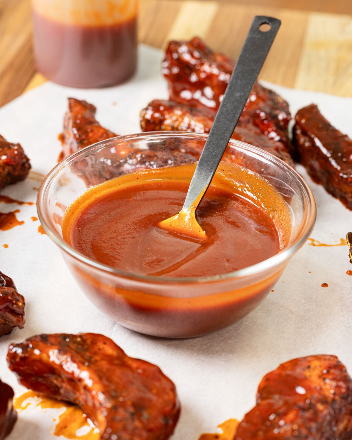 Bowl of buffalo bbq sauce with a spoon, sitting on a cutting board with ribs.
