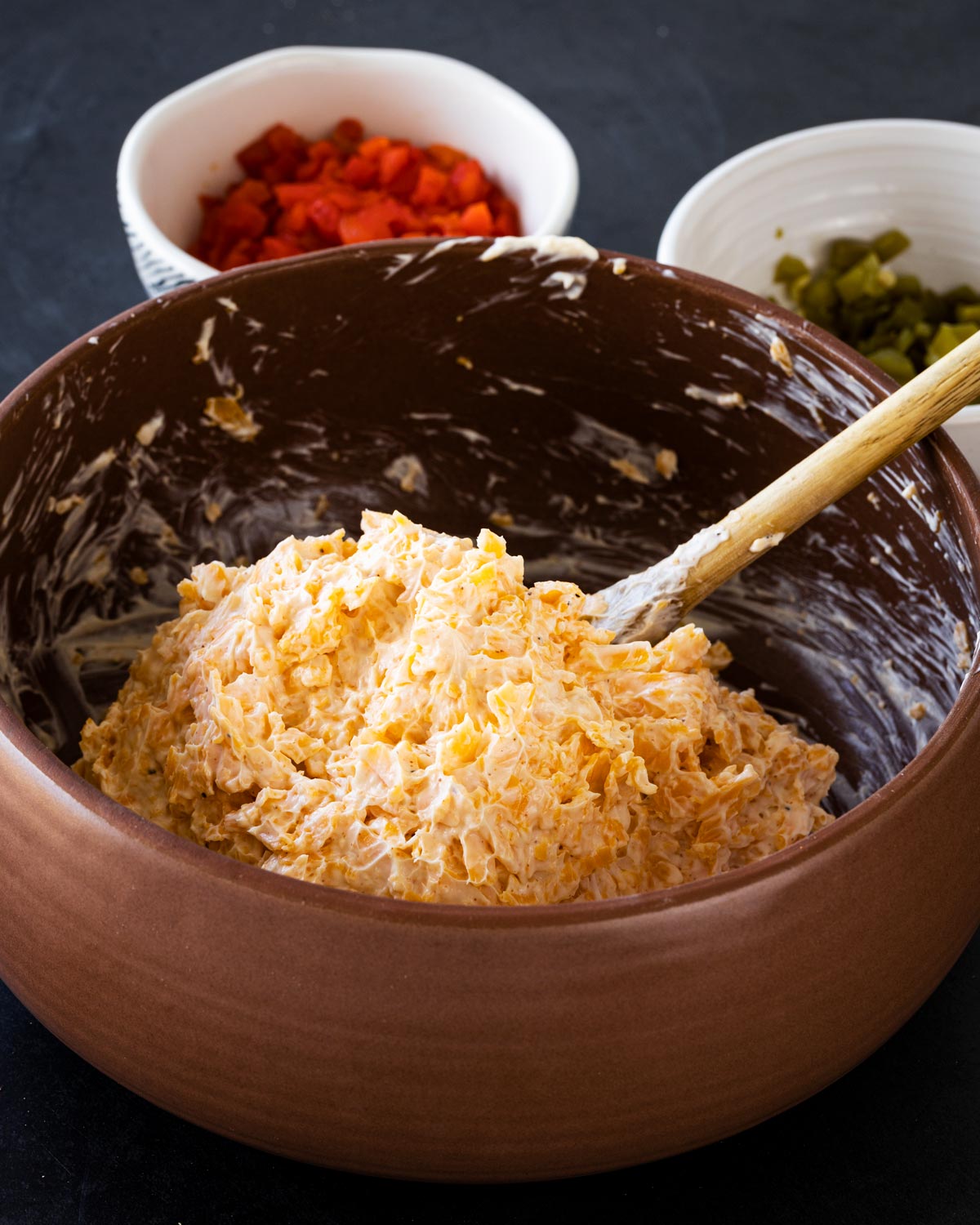 Jalapeño pimento cheese base in a terracotta bowl before peppers are folded in, diced pimentos and pickled jalapeños visible in background bowls