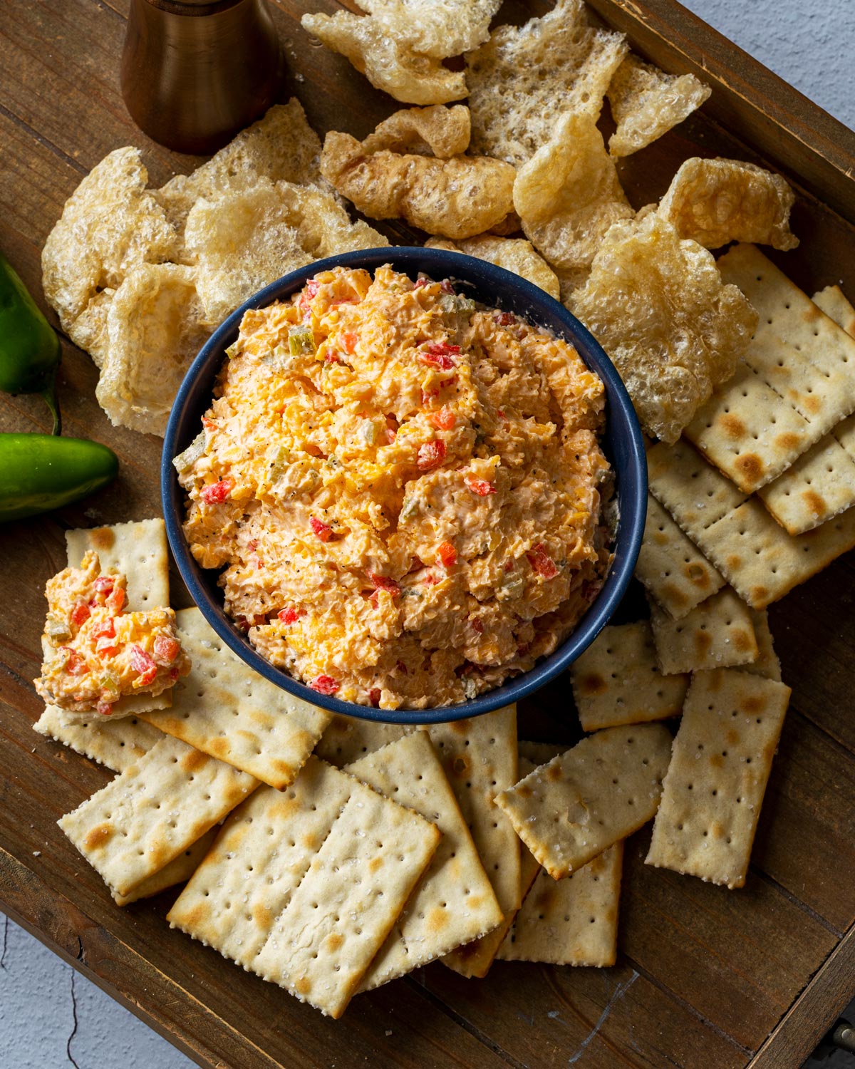 Jalapeño pimento cheese served in a dark bowl surrounded by crackers and pork rinds on a wooden board