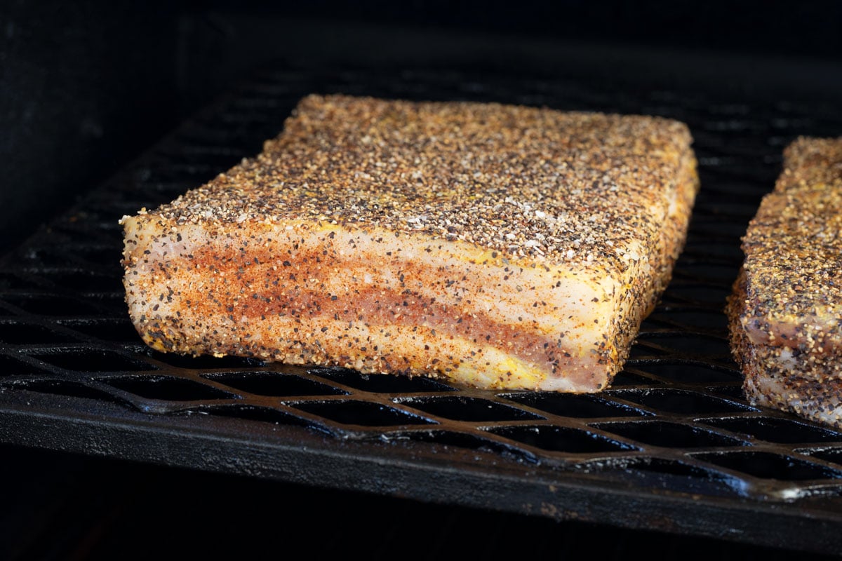 Seasoned pork belly slab fat-side down on smoker grates at the start of the cook