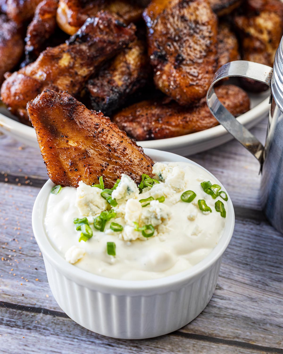 Grilled chicken wing dipped into a ramekin of blue cheese dressing topped with crumbles and scallions, plate of wings behind