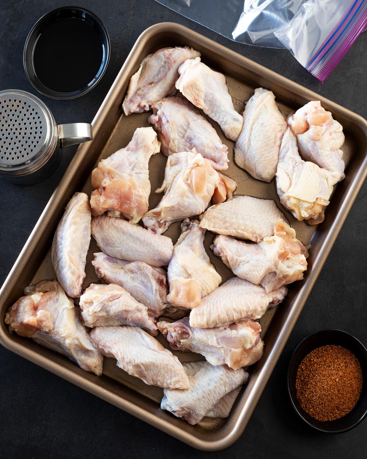 Raw chicken wings spread on a sheet pan next to oil and dry rub before seasoning