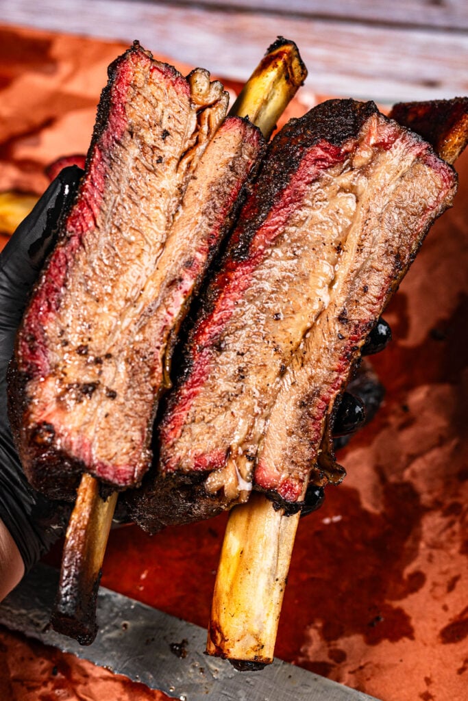 Sliced smoked beef plate ribs held in black gloves showing smoke ring and bark