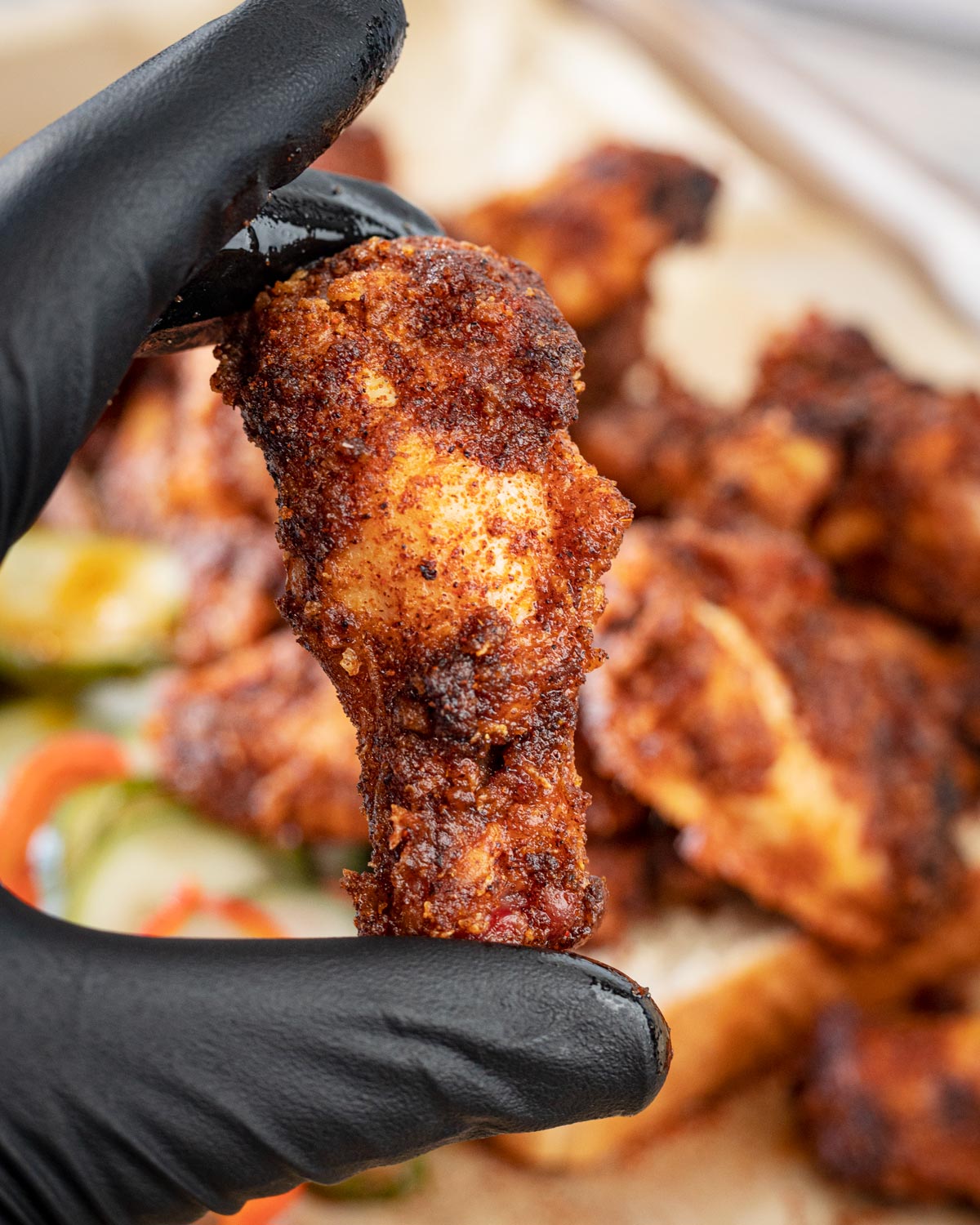 Nashville hot grill fried chicken wing held in black gloves with more wings and pickles in the background