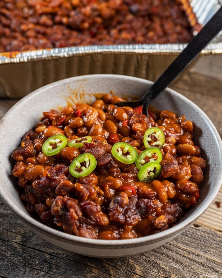 Smoked baked beans with chipotle and bacon served in a bowl with fresh jalapeno slices, foil pan on the smoker in the background