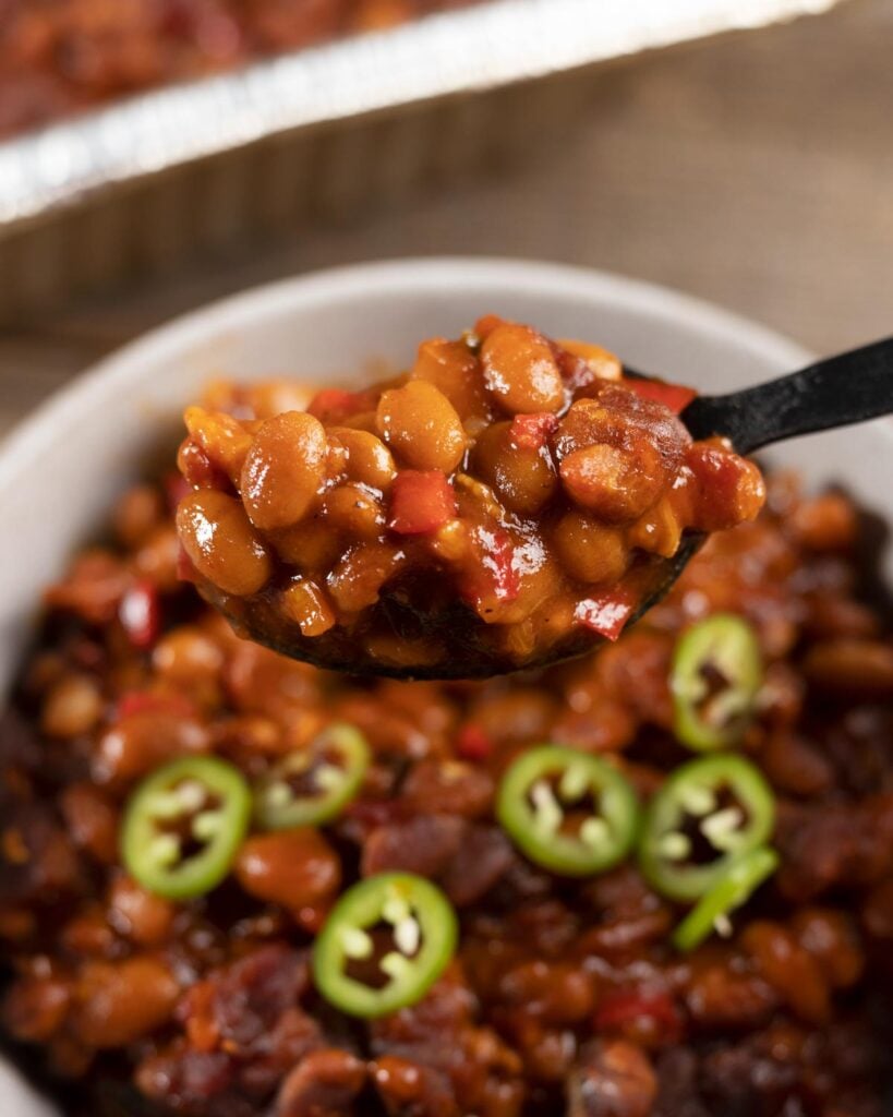 Spoonful of smoked baked beans with chipotle and bacon lifted from a bowl garnished with fresh jalapeno slices