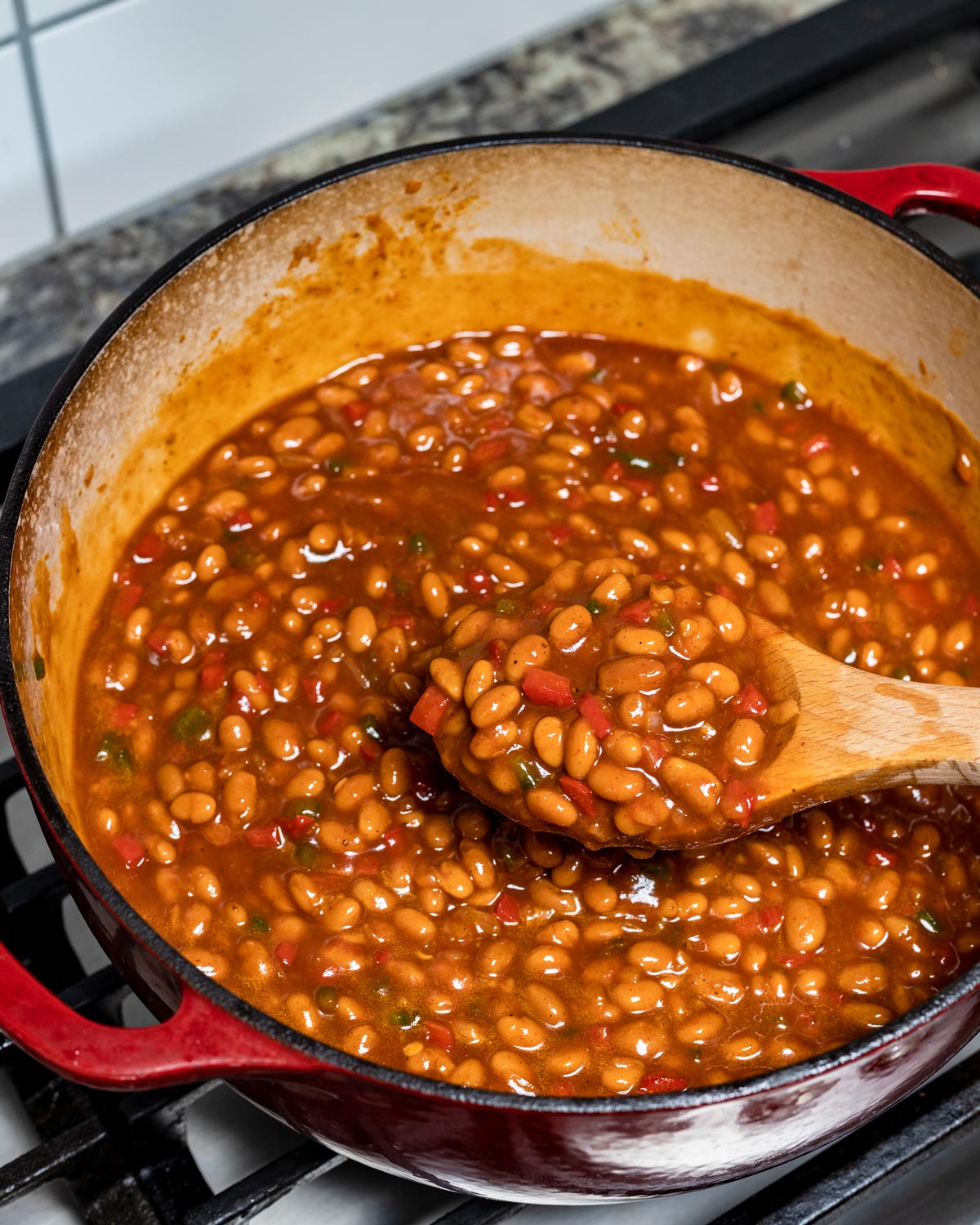 Canned baked beans combined with BBQ sauce and aromatics simmering in a red Dutch oven before smoking