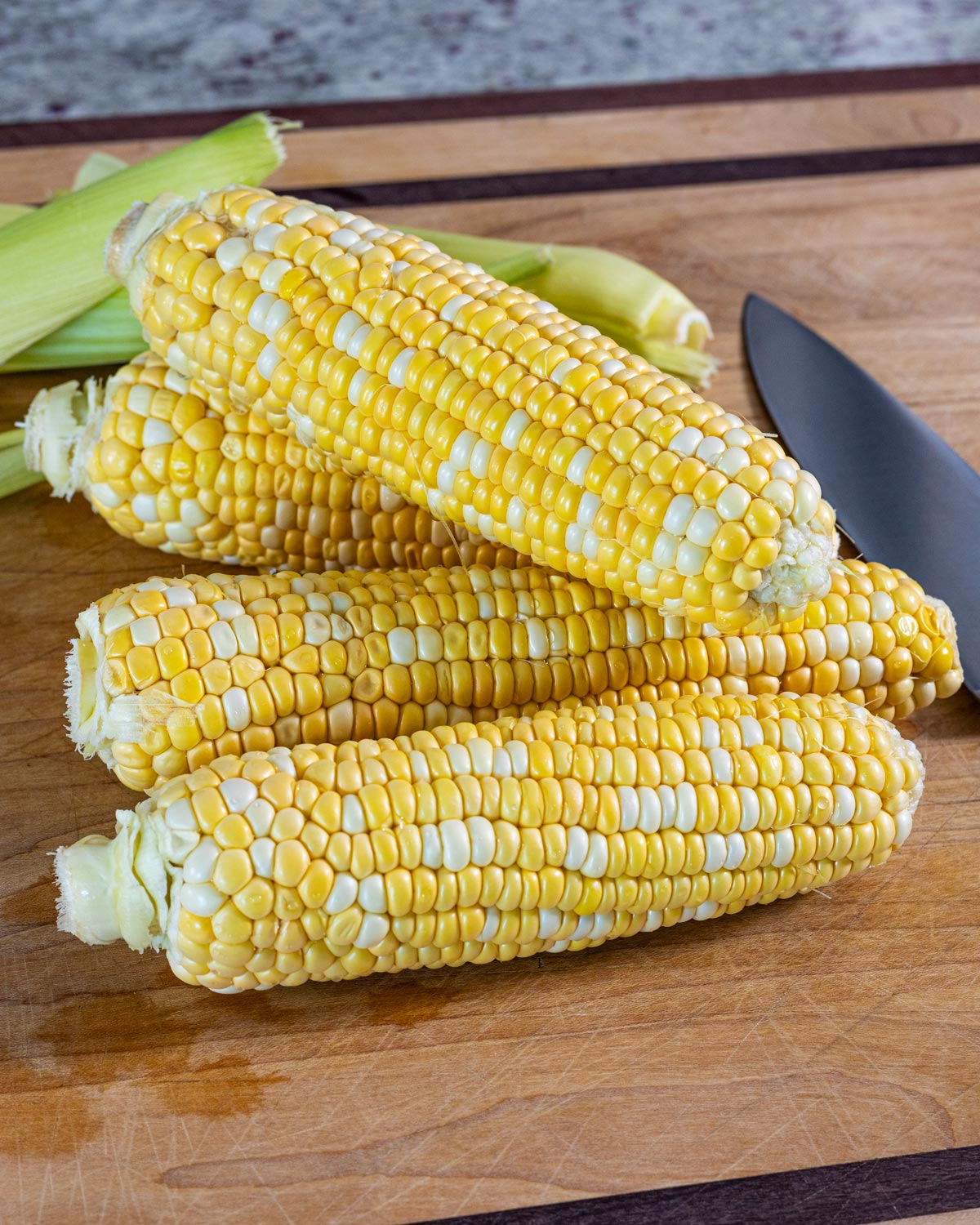 Fresh husked corn cobs on a cutting board with a knife, ready to be cut into corn ribs