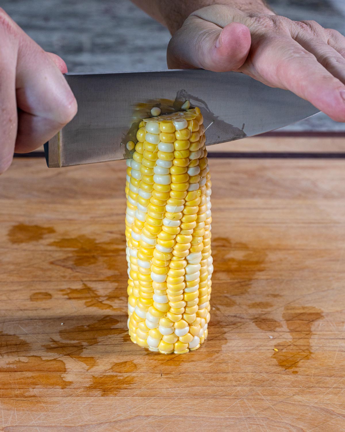 Two hands pressing a knife through the top of an upright corn cob to slice into corn ribs