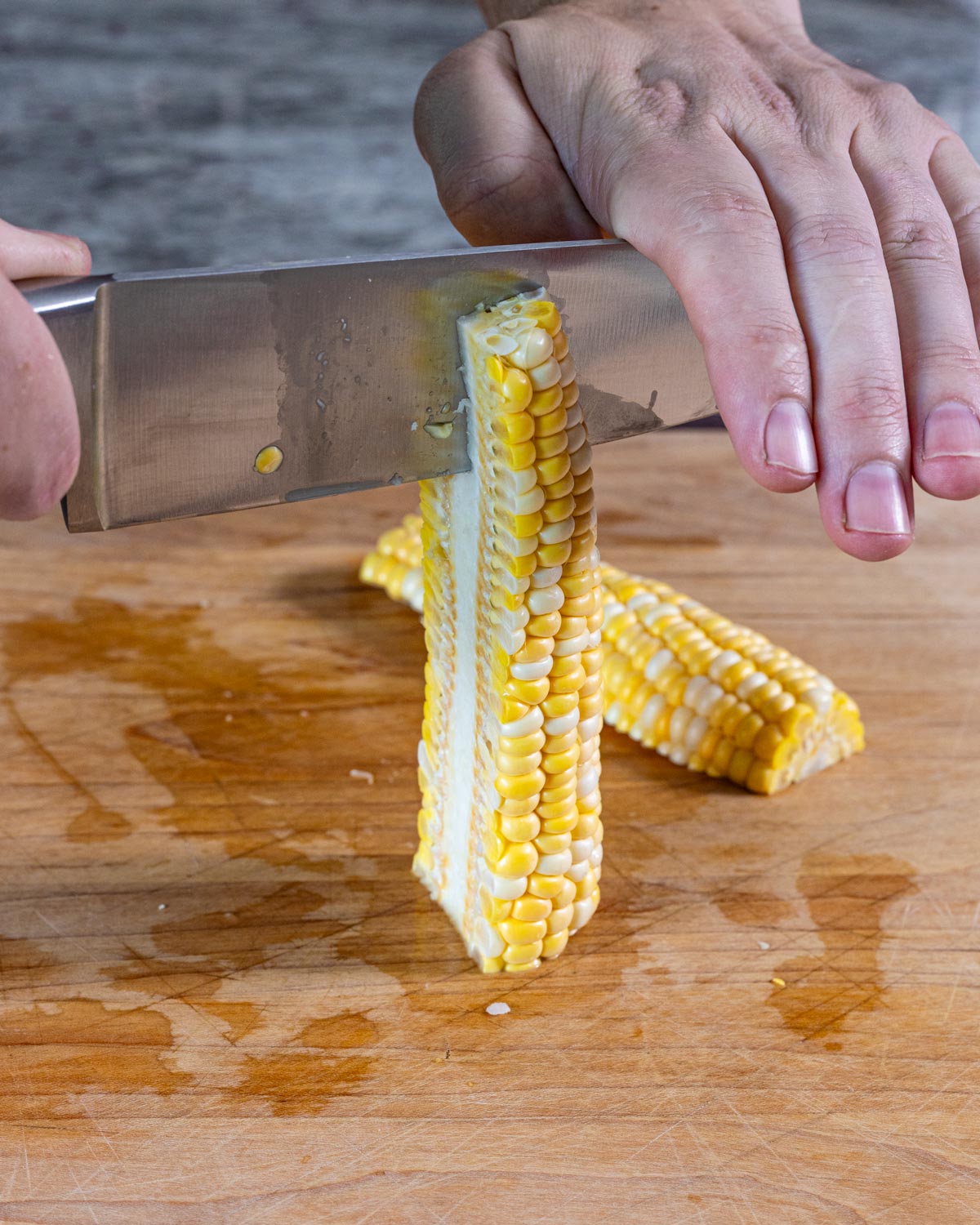 Hands pressing a knife through a corn cob standing upright to quarter it into corn ribs on a cutting board