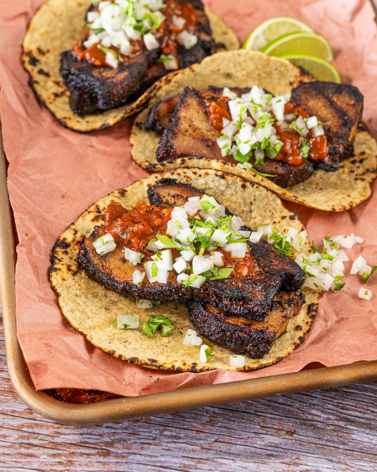 Grilled pork belly tacos on corn tortillas with red salsa and white onion on a sheet pan, lime wedges alongside