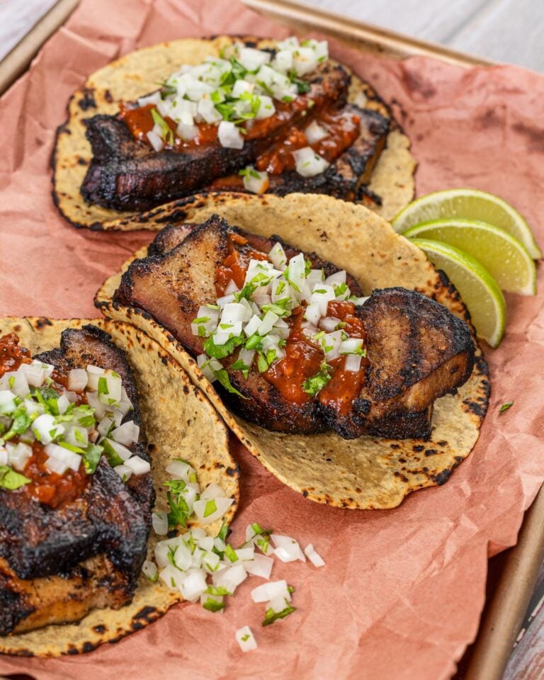 Grilled pork belly tacos on charred corn tortillas with red salsa, diced onion, and lime wedges on butcher paper
