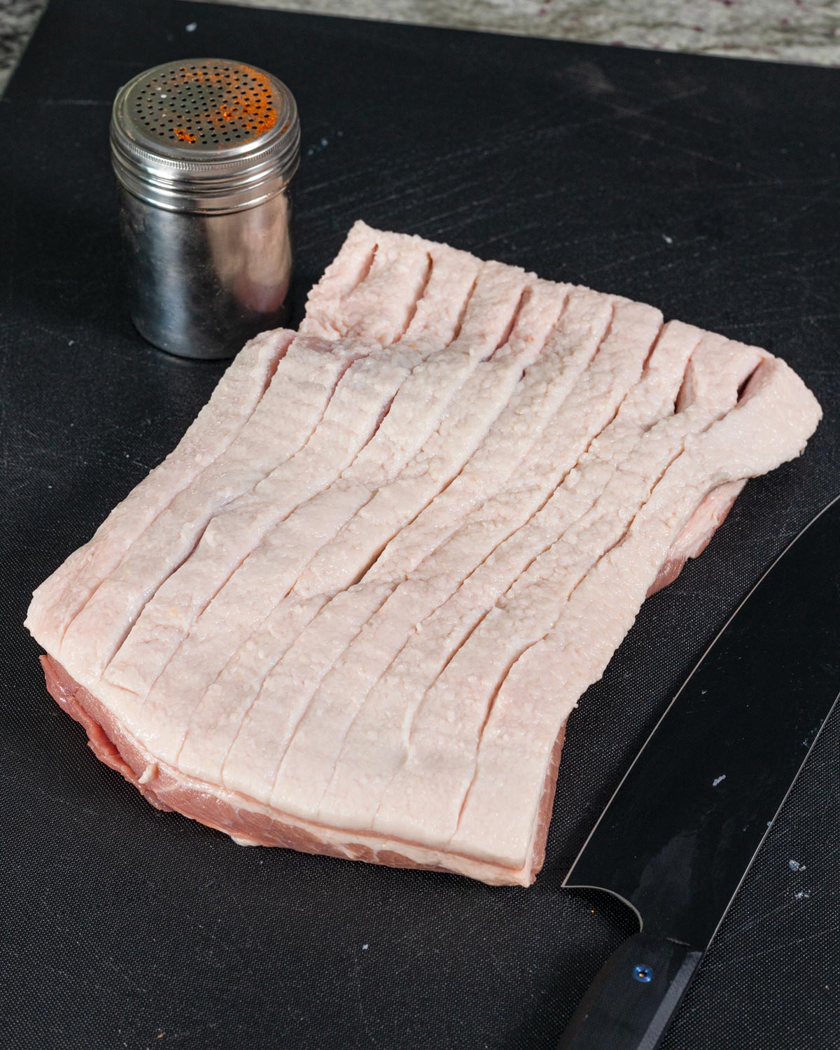 Raw scored pork belly slab fat side up on a cutting board with a seasoning shaker and knife alongside
