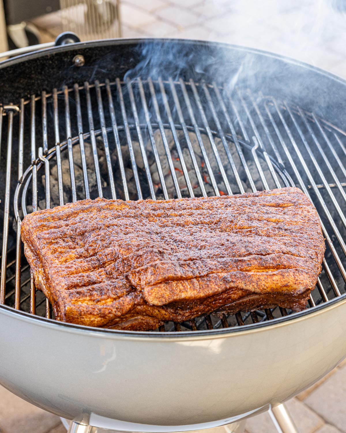 Seasoned pork belly slab placed fat side up on a charcoal grill for indirect smoking, smoke rising from the coals