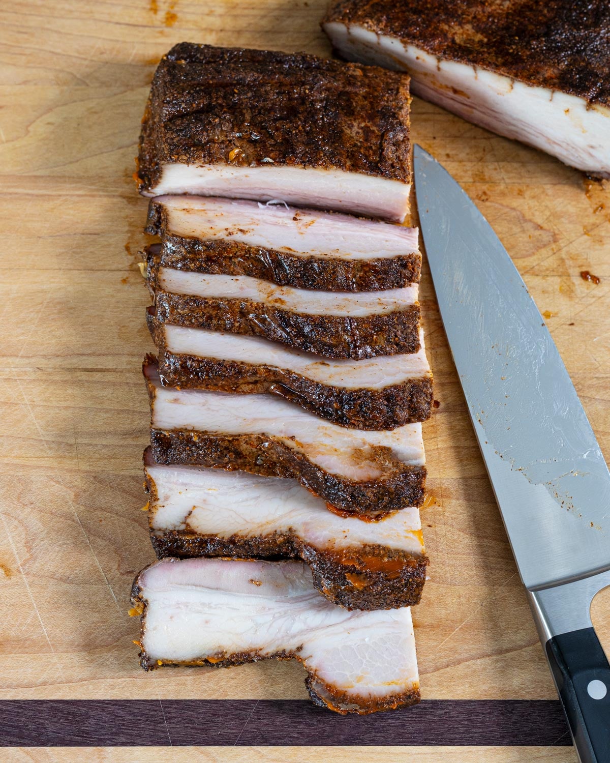 Smoked pork belly sliced into strips on a cutting board showing the meat and fat layers, chef's knife alongside