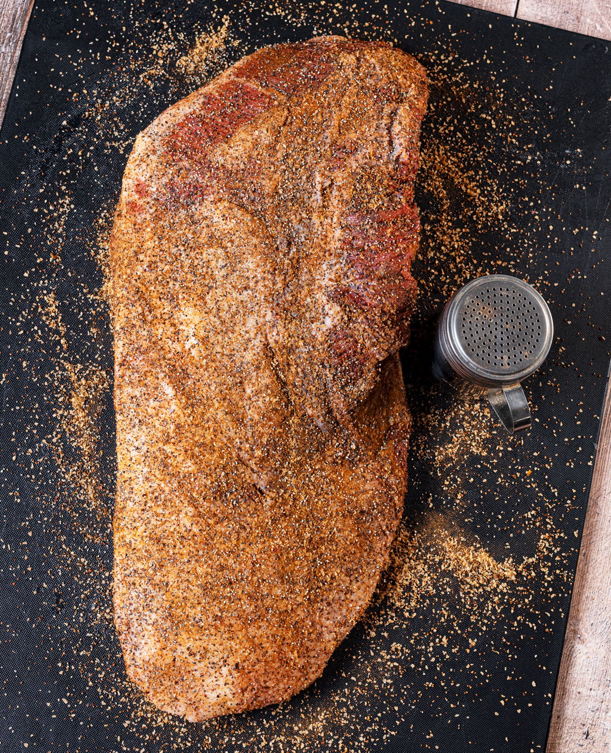 Whole packer brisket generously seasoned with beef rub on a black cutting mat with a seasoning shaker