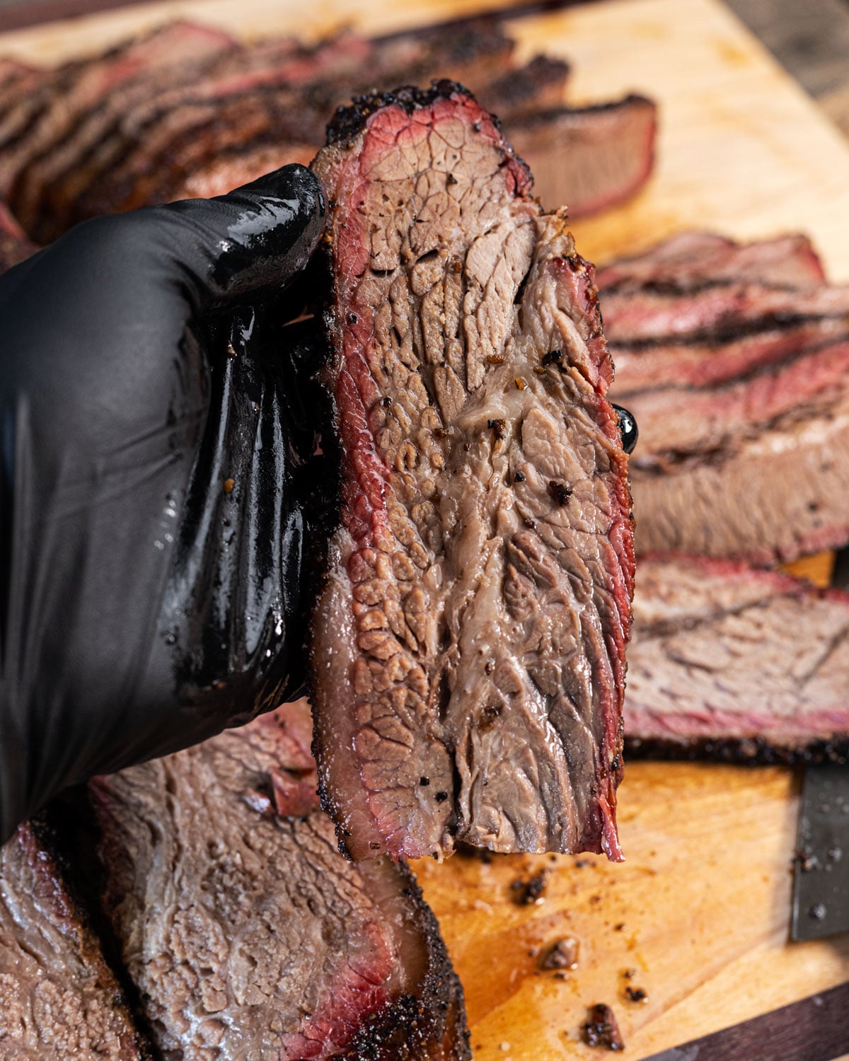 Close up of a smoked brisket flat slice held in a black glove showing smoke ring and grain texture