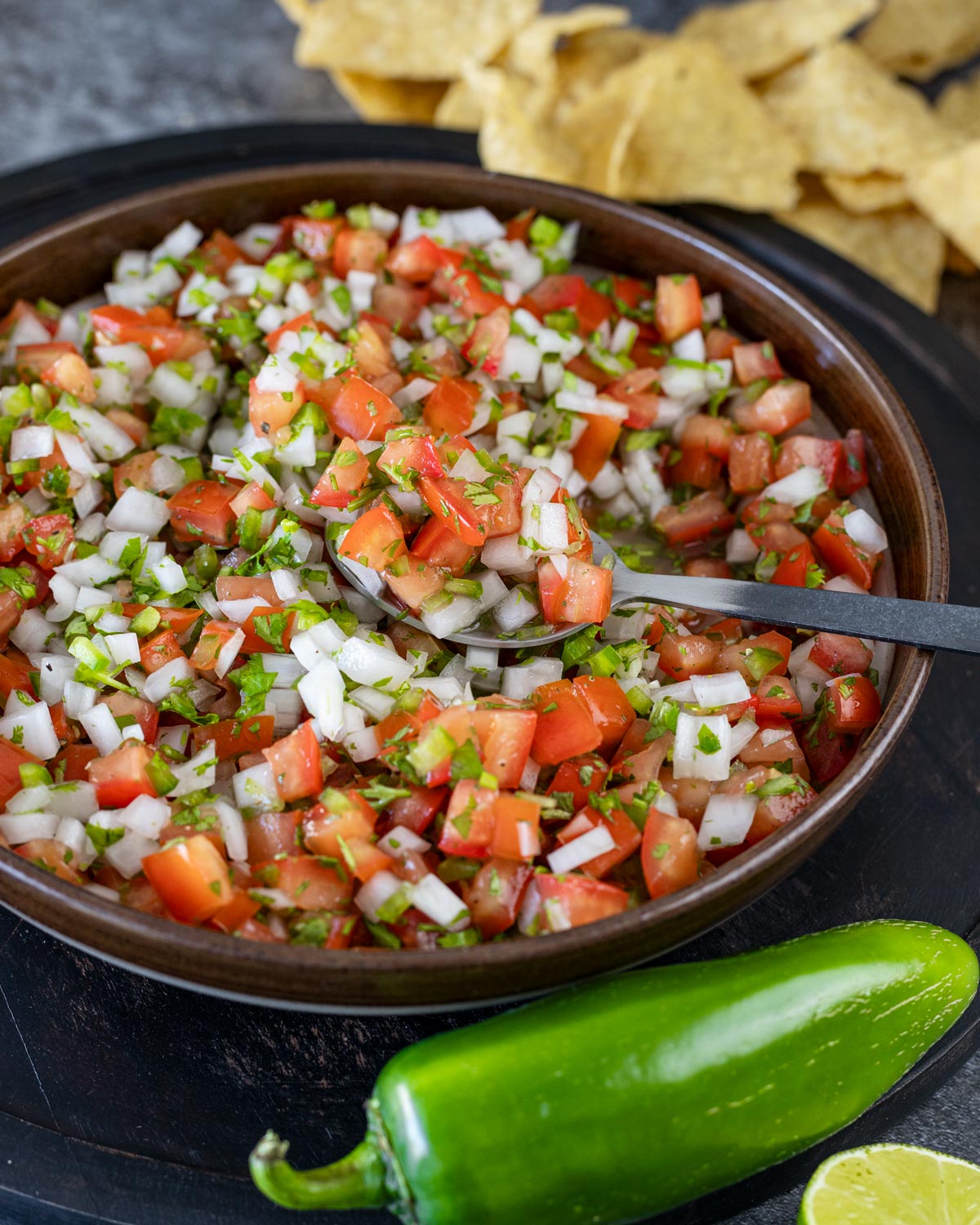 fresh pico de gallo in a wooden bowl with a spoon, jalapeño and lime wedge alongside