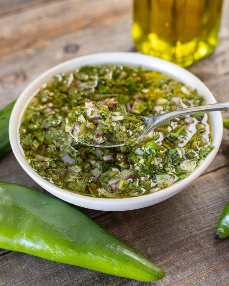 Spoonful of Hatch chile chimichurri lifted from a white bowl, fresh green chile and olive oil behind