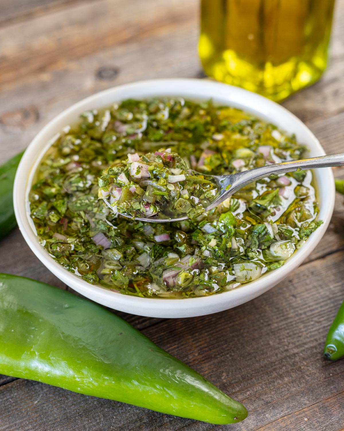 Spoonful of Hatch chile chimichurri lifted from a white bowl, fresh green chile and olive oil behind
