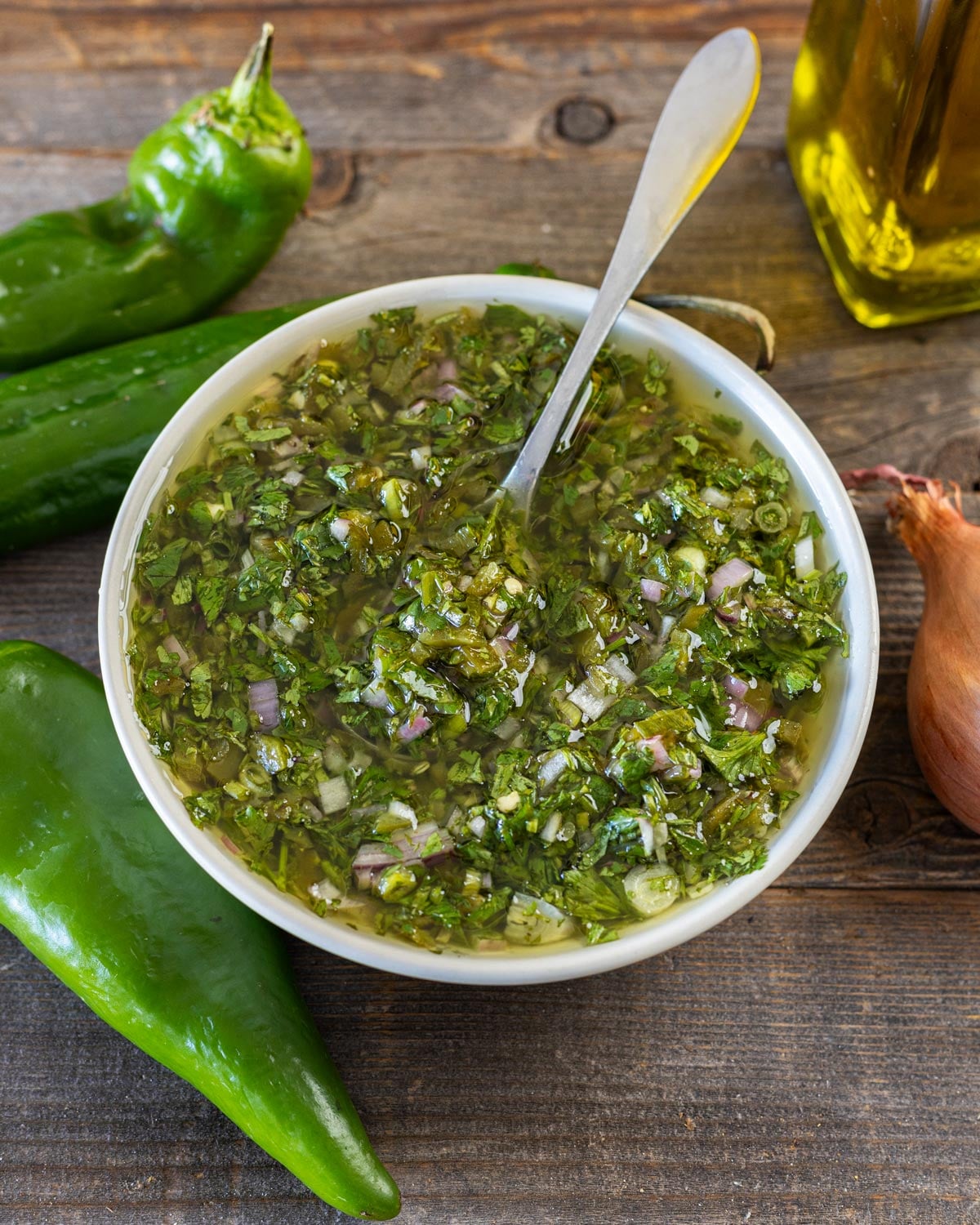 Hatch chile chimichurri in a white bowl with a spoon, whole green chiles and chopped roasted chiles behind