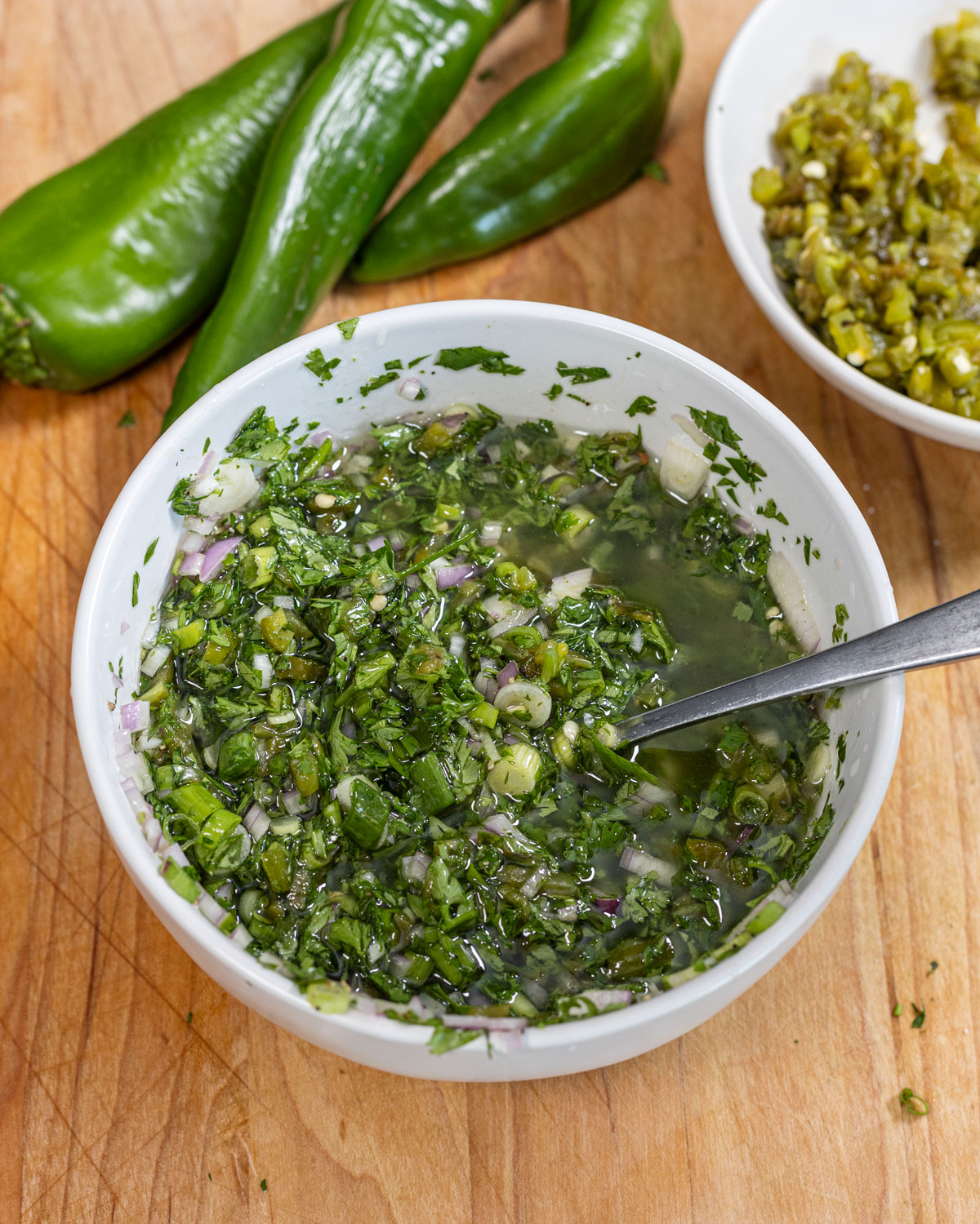 Hatch chile chimichurri in a white bowl with a spoon, whole green chiles and a dish of chopped roasted chiles behind