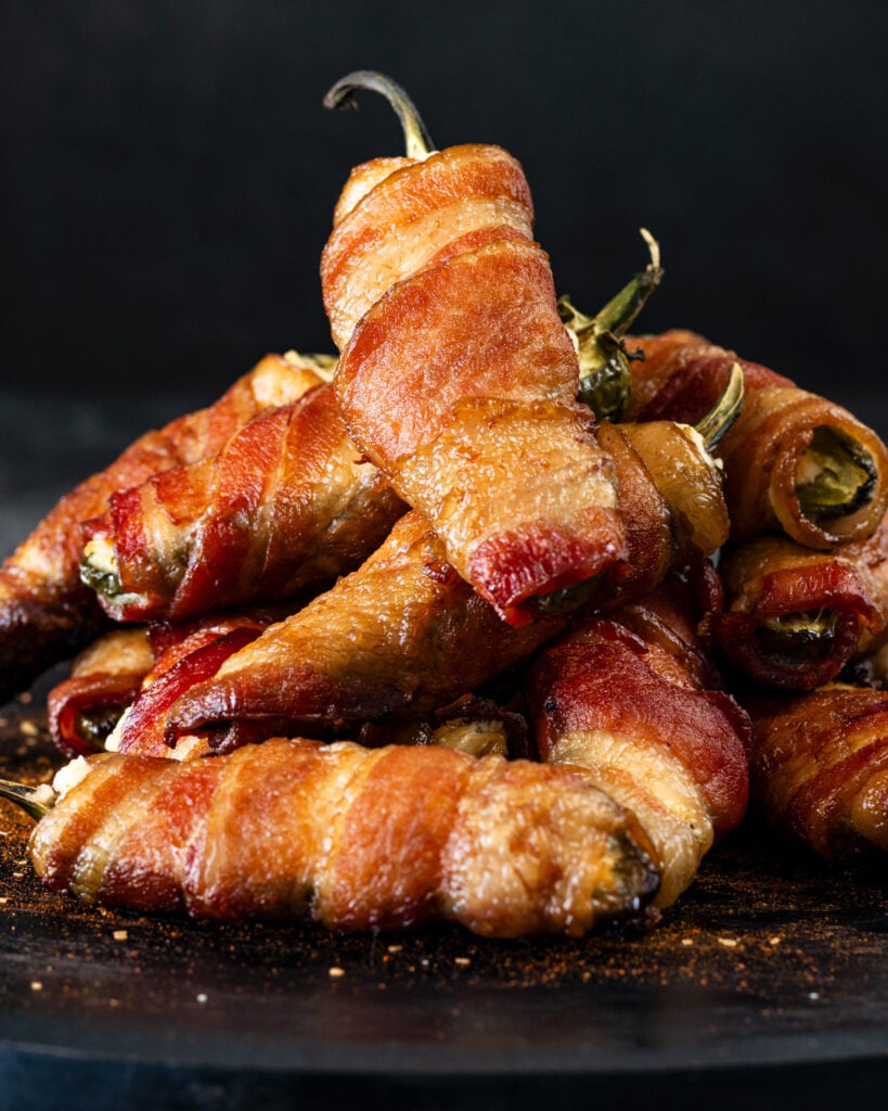 Bacon-wrapped smoked jalapeño poppers stacked with BBQ rub dusted around the base, dark background