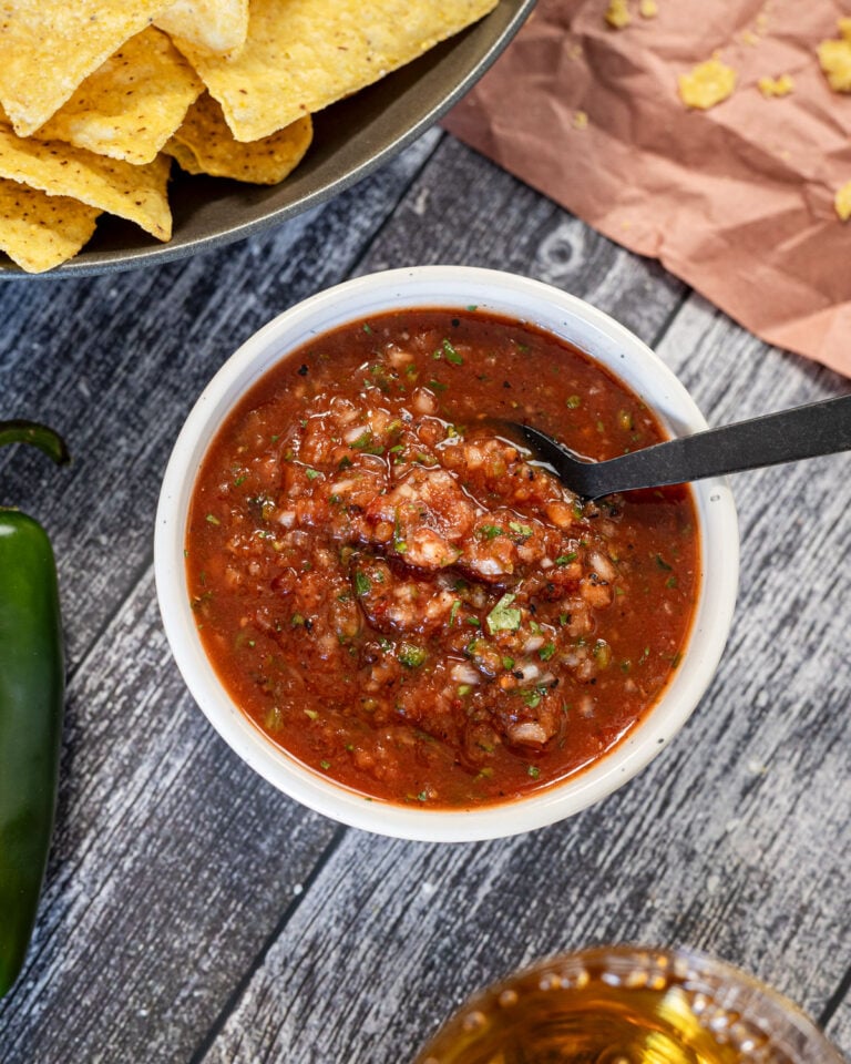 Restaurant-style chipotle salsa in a white bowl with a spoon, tortilla chips, fresh jalapeño, and a cold beer