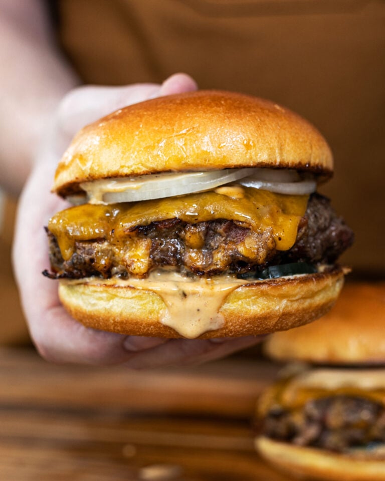 Crack burger held up showing melted cheddar, BBQ ranch sauce, and raw onion on a toasted brioche bun