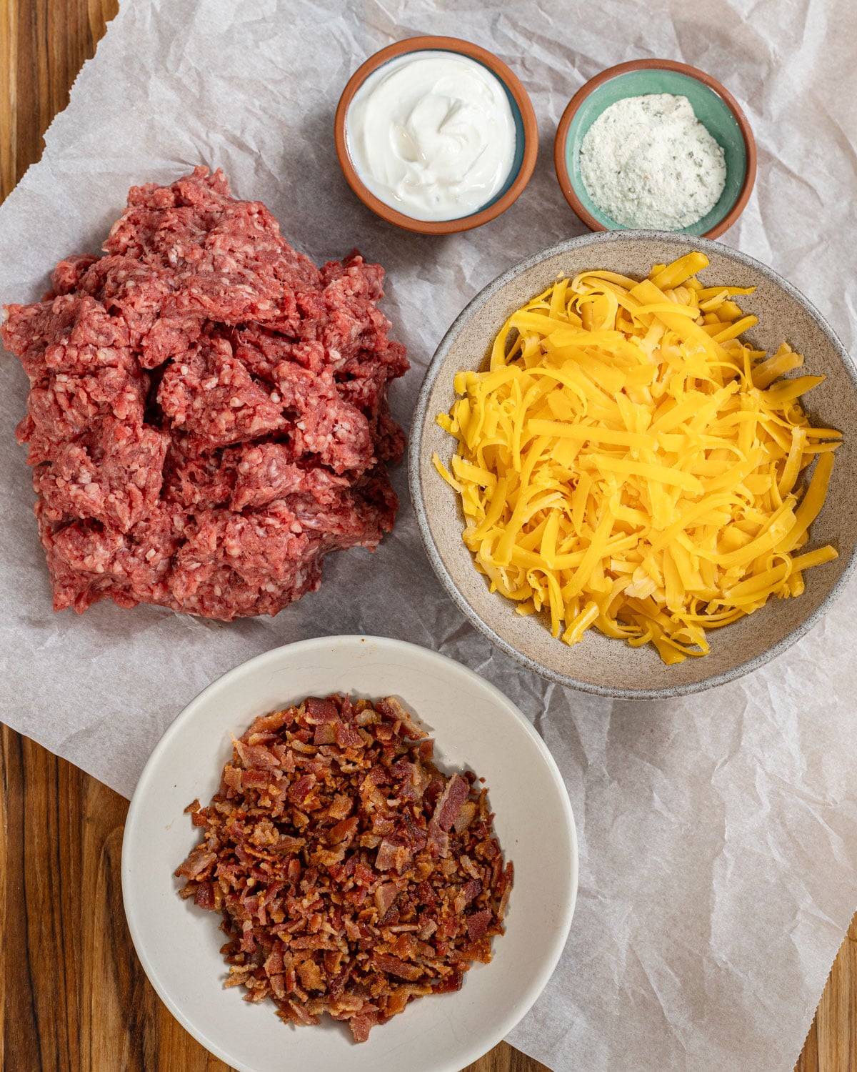 Crack burger ingredients laid out on parchment: ground beef, shredded cheddar, crumbled bacon, sour cream, and ranch powder