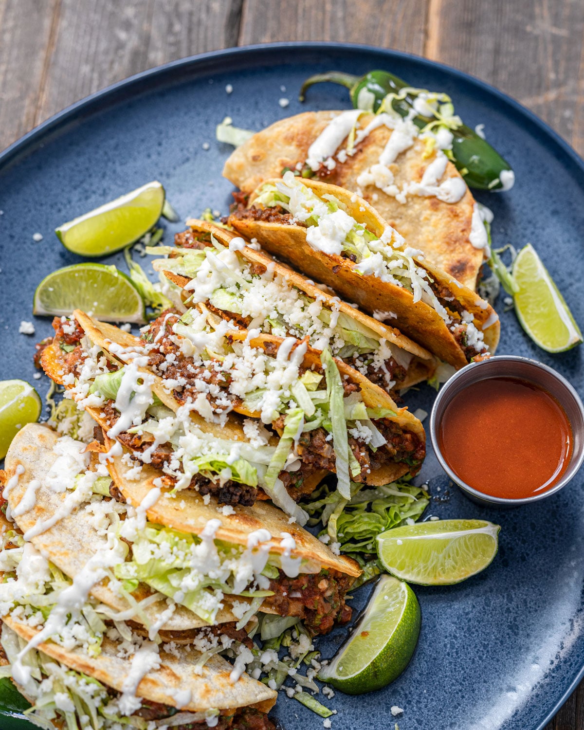 Plated tacos dorados topped with shredded iceberg, cotija cheese, and Mexican crema, served with red salsa and lime wedges