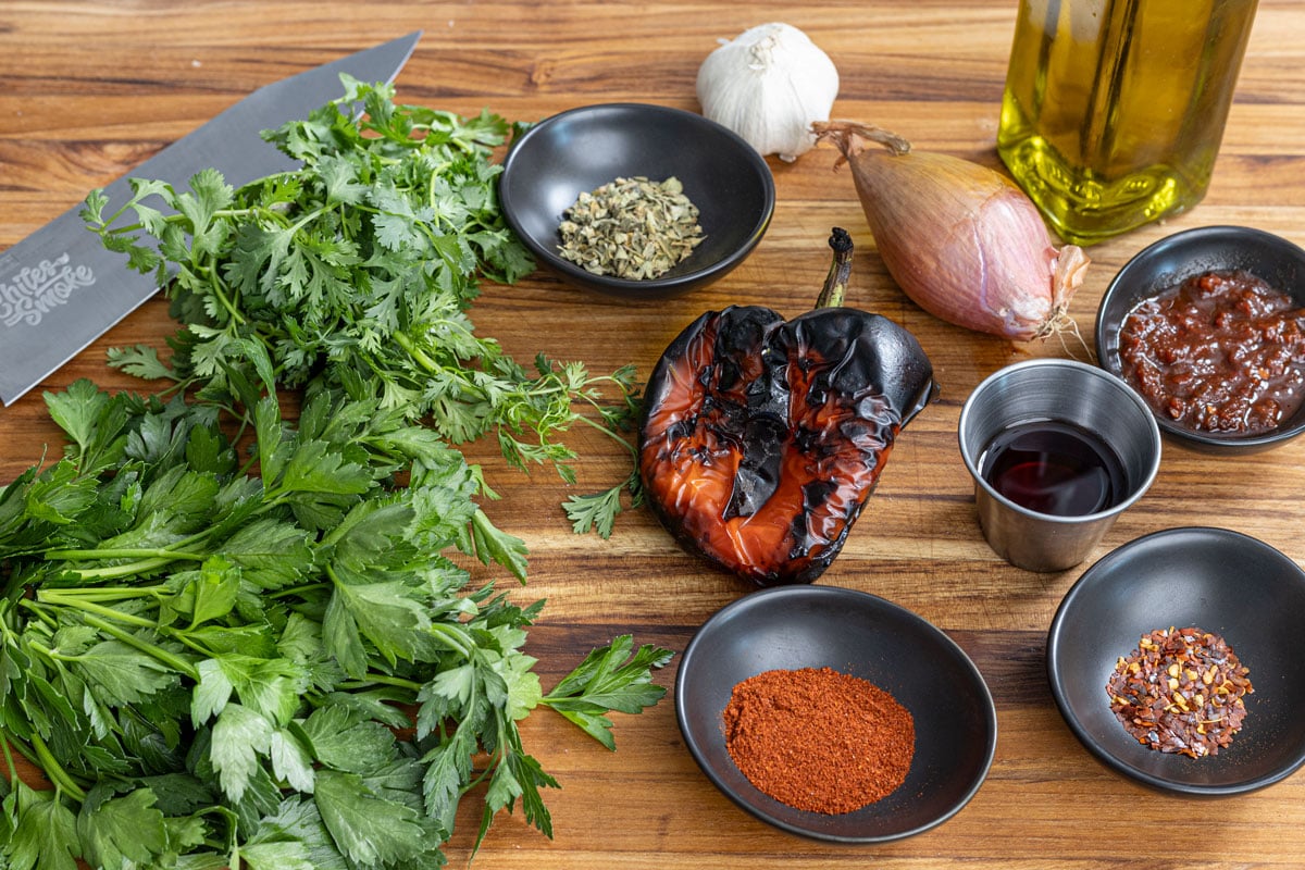 Ingredients for red chimichurri on a cutting board: parsley, cilantro, a charred red pepper, shallot, garlic, chipotles in adobo, smoked paprika, red pepper flakes, and red wine vinegar