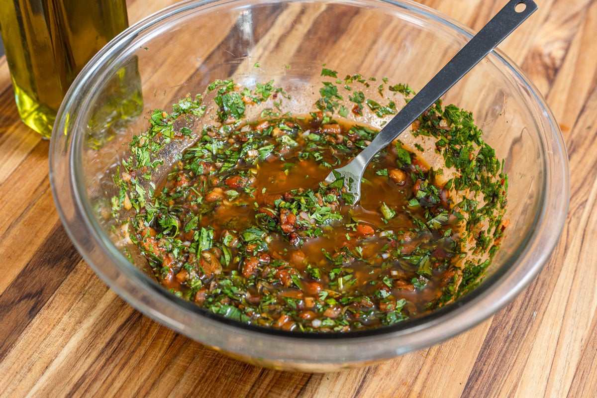 Red chimichurri freshly mixed in a glass bowl with a fork, olive oil bottle behind