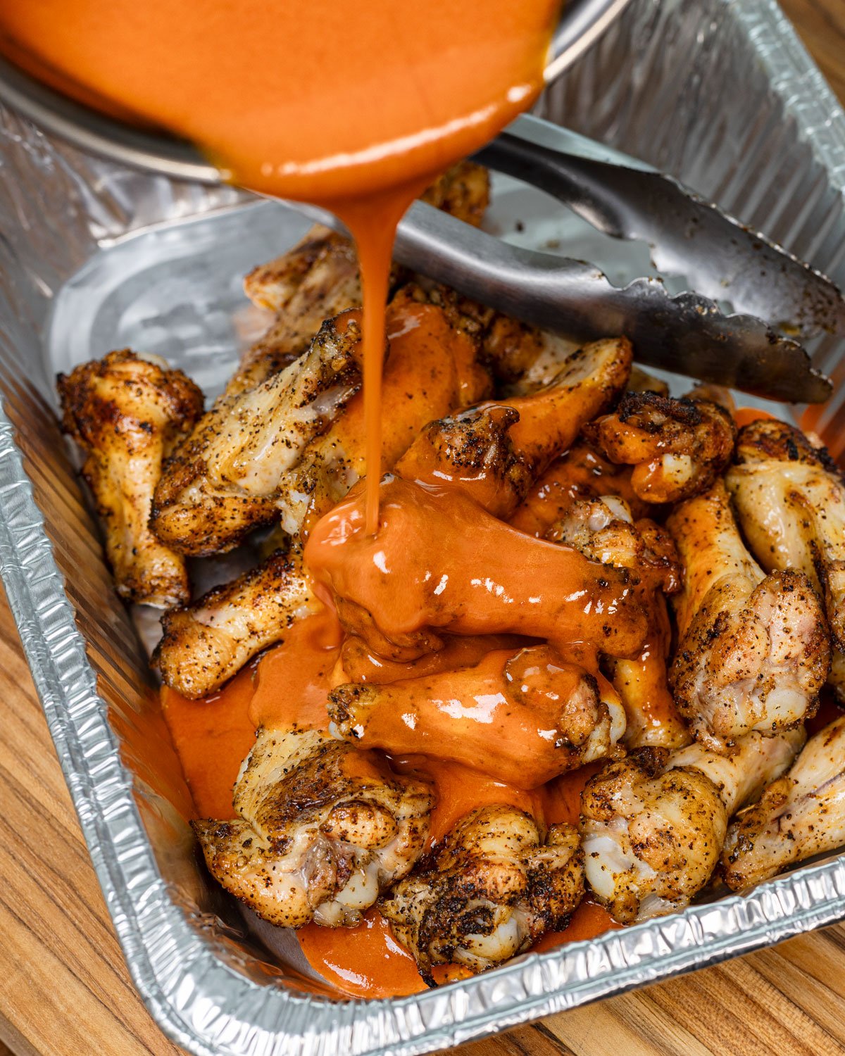 Wings in a foil pan with buffalo sauce being poured over the top.
