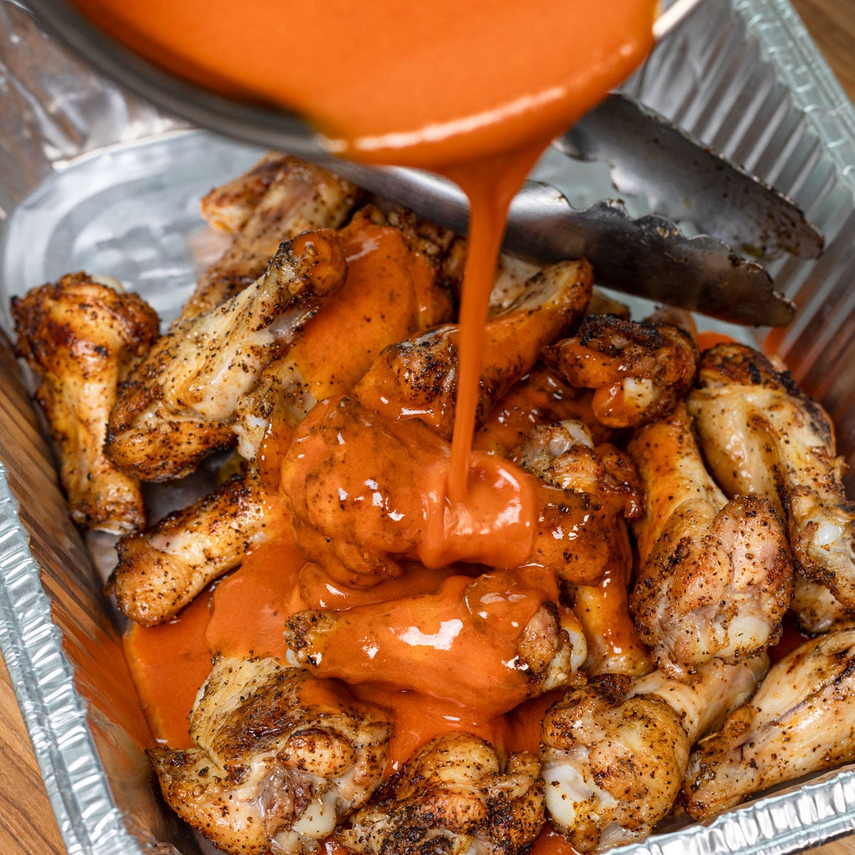 Creamy buffalo sauce poured over grilled chicken wings in a foil pan
