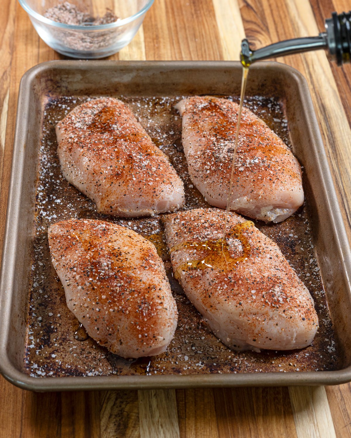 Four chicken breasts on a sheet pan being seasoned and drizzled with olive oil.