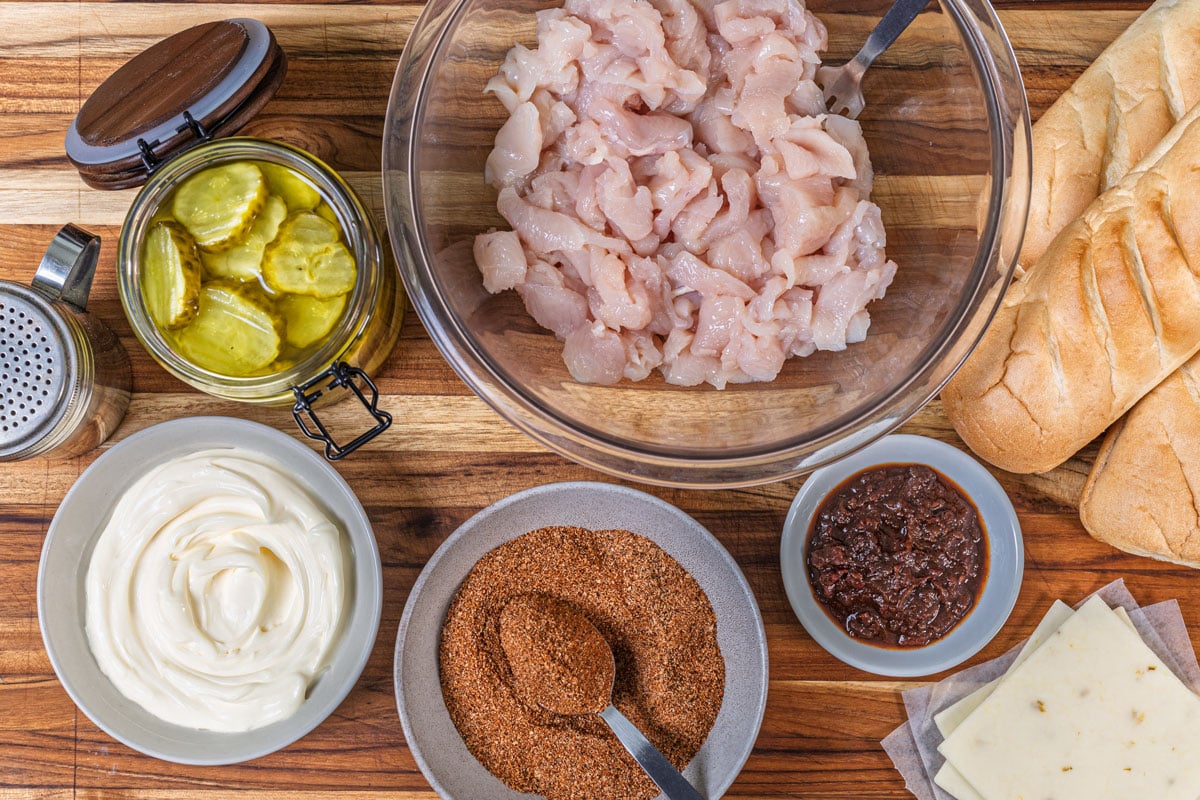 Nashville hot chicken melt ingredients on a wood cutting board, including raw sliced chicken breast, seasoning, pickles, mayo, pepper jack, and hoagie rolls