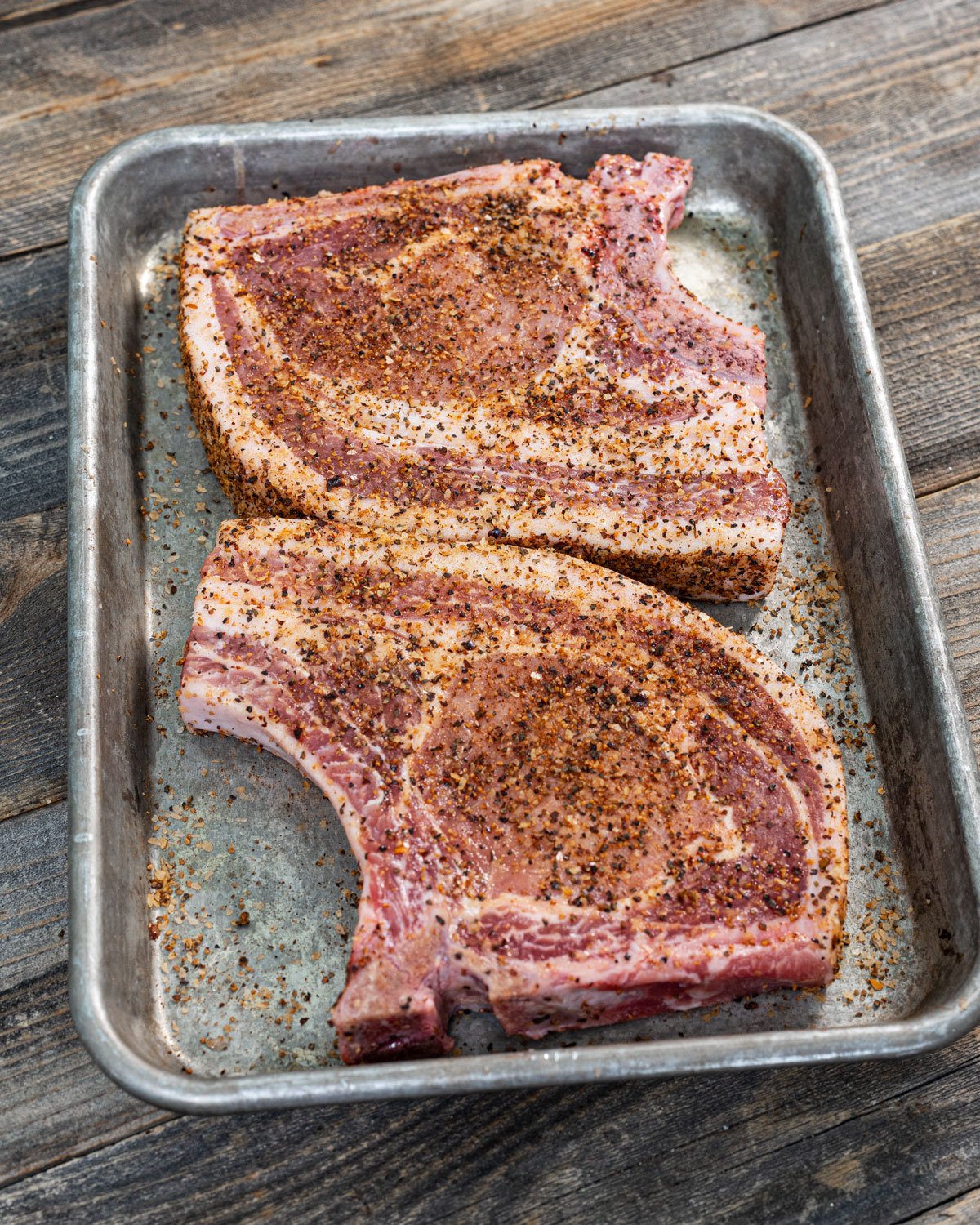 Two bone-in pork chops on a sheet pan with seasoning on them.