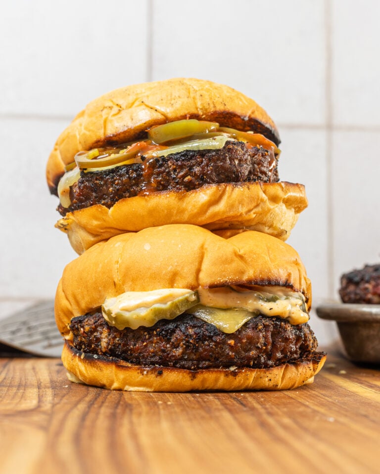 Two charcoal burgers stacked on a wooden cutting board with melted cheese, pickles, and toasted brioche buns