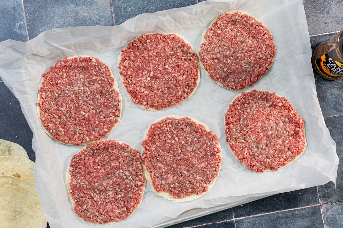 Ground beef pressed into tortillas on a baking sheet for tacos