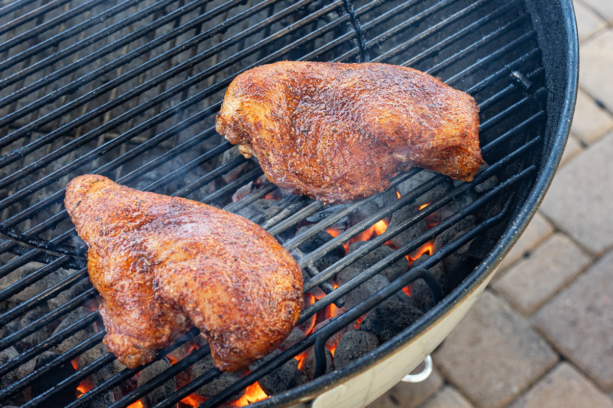 Two seasoned chicken breasts searing over live charcoal on a kettle grill