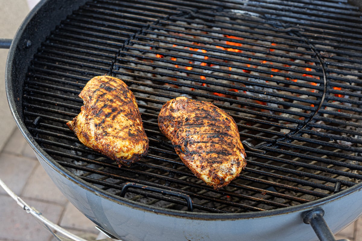 Two grilled chicken breasts with defined grill marks resting on the indirect side of a charcoal kettle grill