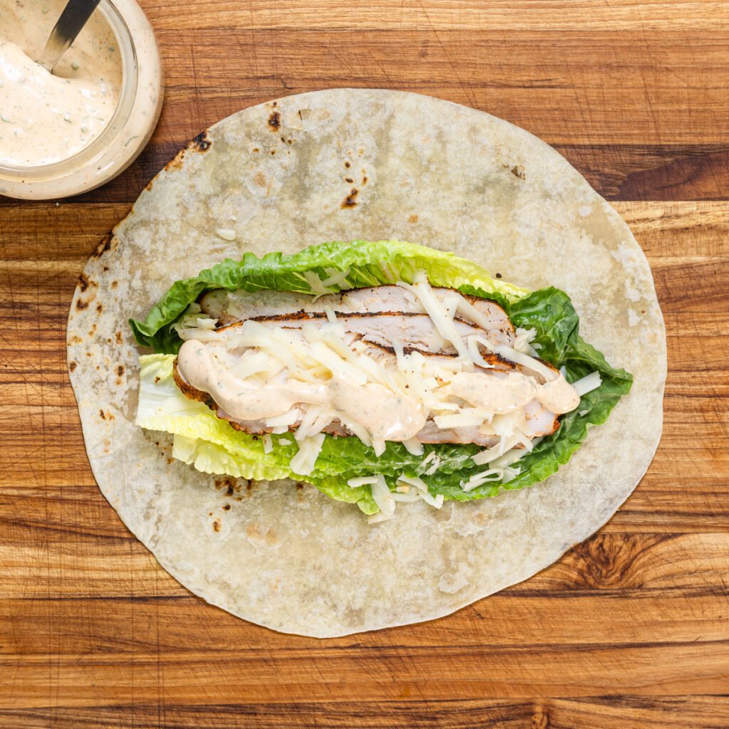 Assembling the grilled chicken wrap with lettuce, chicken, cheese and sauce.