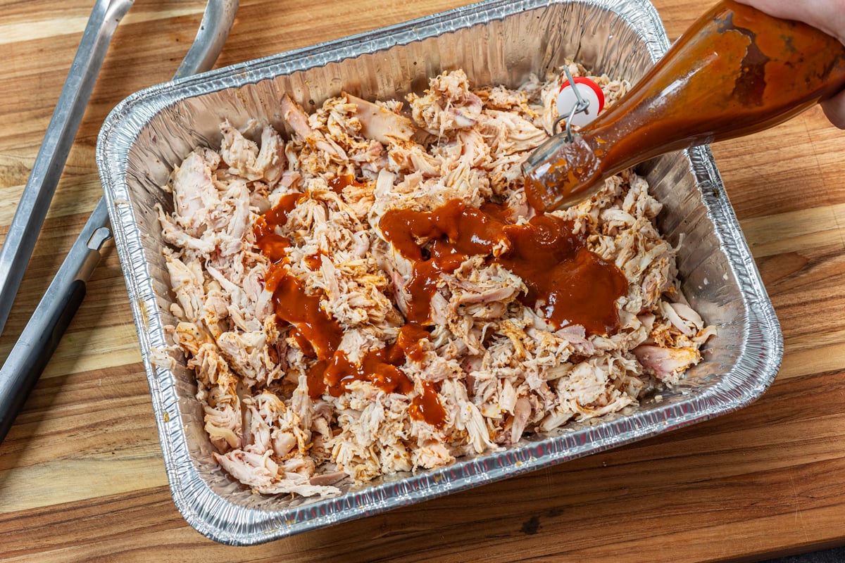 Pouring BBQ sauce onto the smoked pulled chicken in a foil pan.