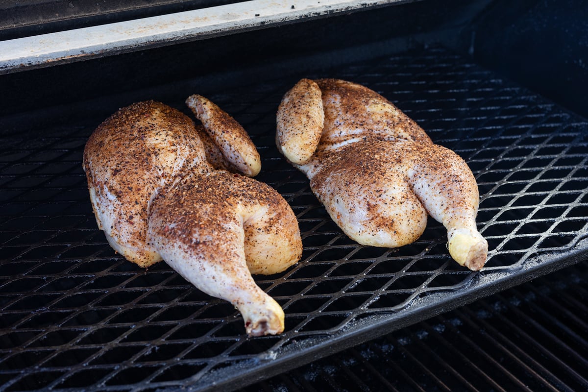 Two seasoned chicken halves placed on the grill grates to smoke.