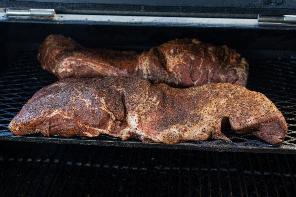 Two butterflied pork butts laid flat and open on smoker grates coated in dry rub at the start of the cook