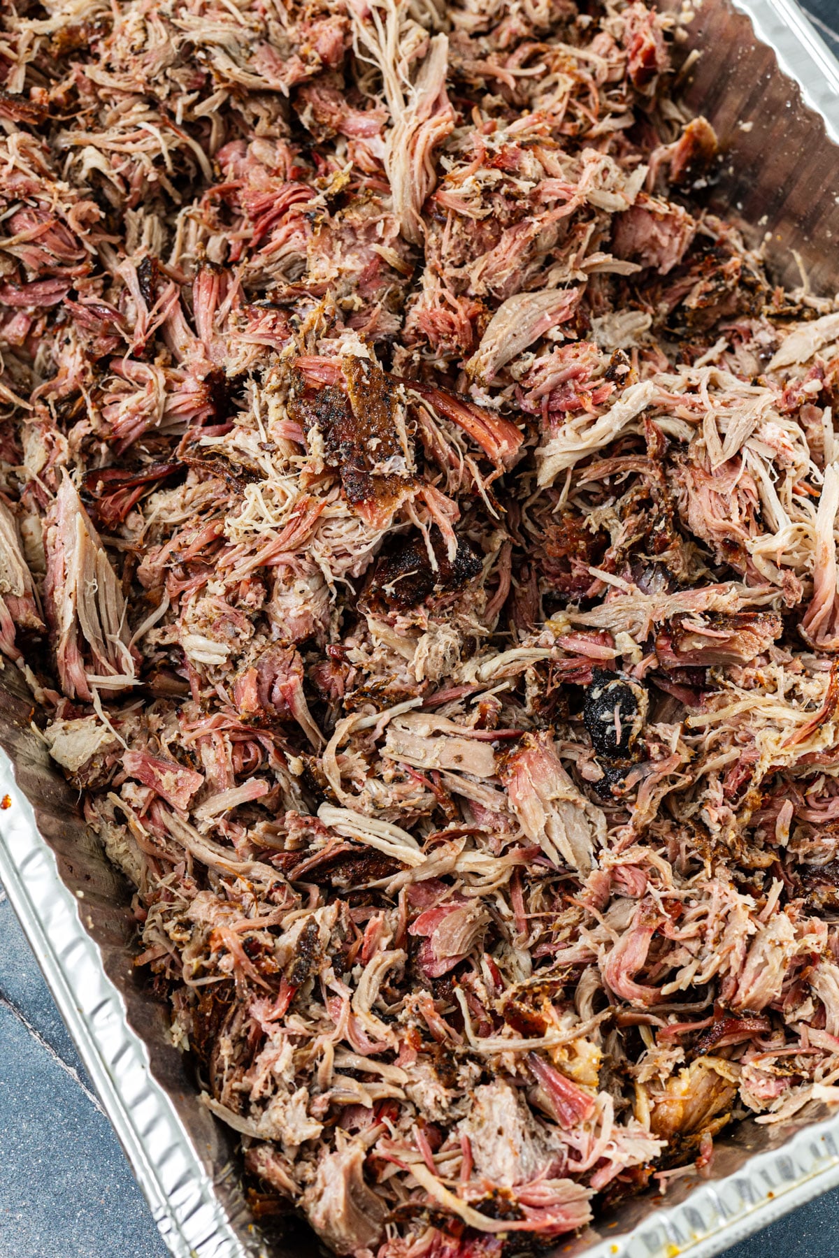 Fully shredded smoked butterflied pork butt with dark bark pieces mixed throughout in an aluminum pan