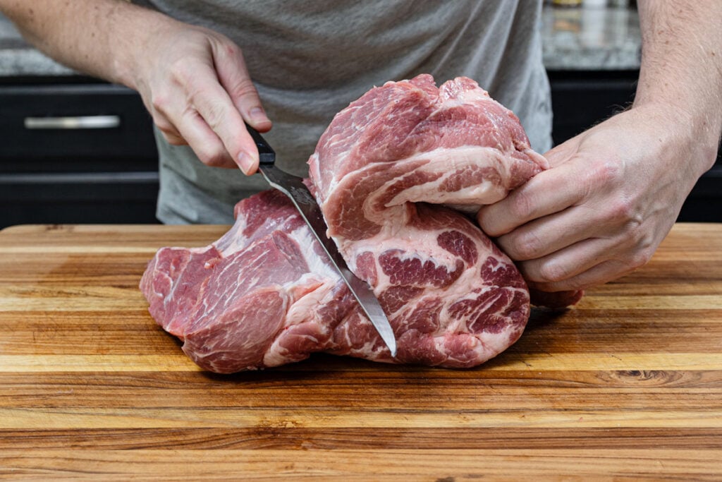 Knife following the natural muscle seams of a pork butt to butterfly it open on a wood cutting board