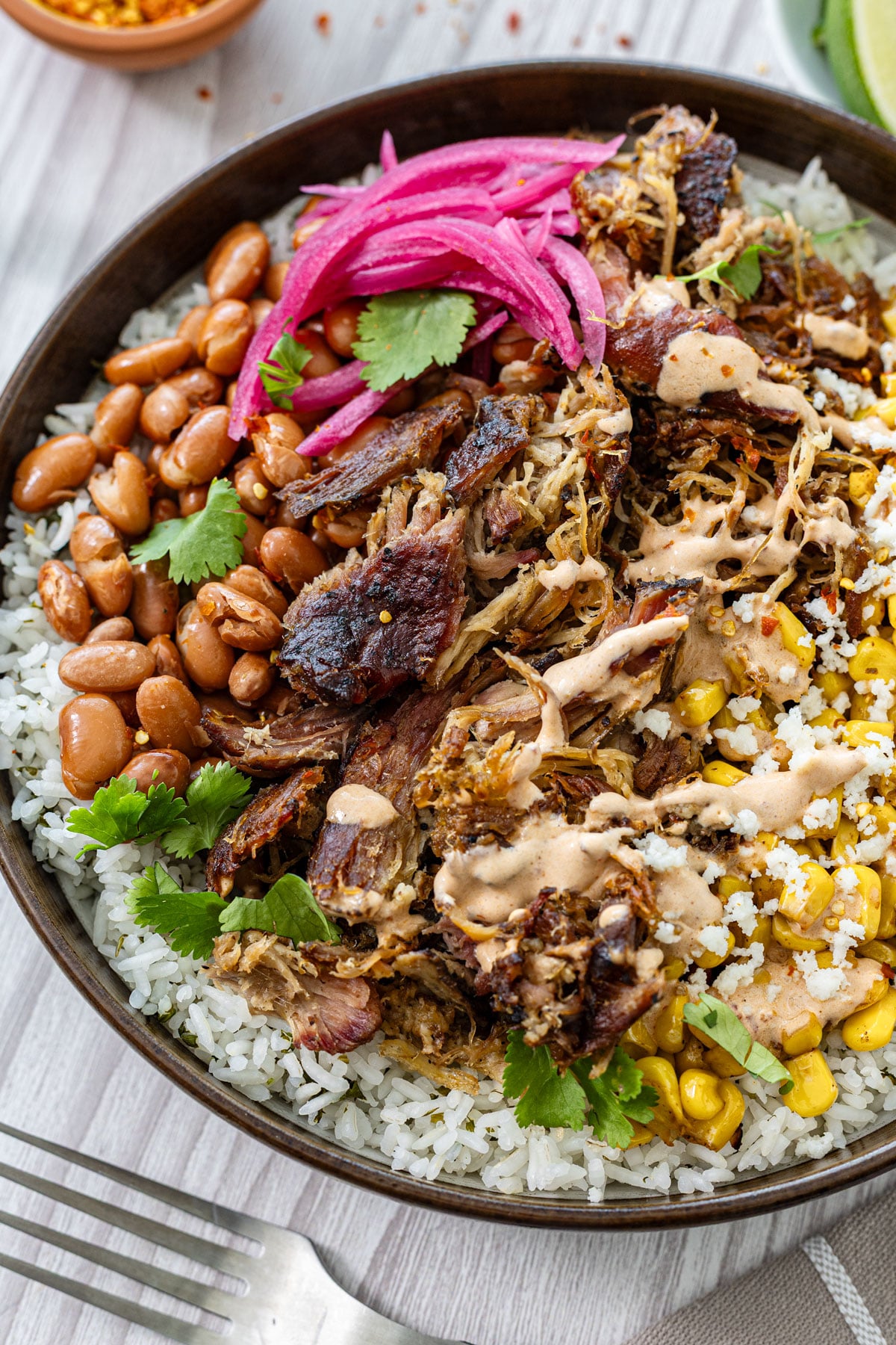 Close up of smoked carnitas burrito bowl showing crisped pork edges, chipotle lime crema drizzle, and cotija over cilantro lime rice